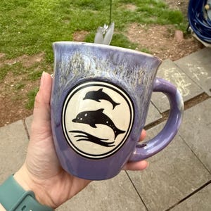 Può includere: Una tazza in ceramica viola con un designo circolare bianco con due delfini neri. La tazza ha una glassa macchiata e un manico bianco.
