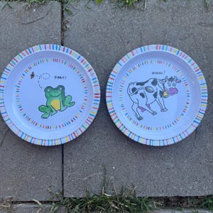 Può includere: Due piatti bianchi con bordi a righe colorate. Uno presenta una rana verde con la scritta "Ribbit!". L'altro ha un'illustrazione di mucca con la scritta "Moooo!".