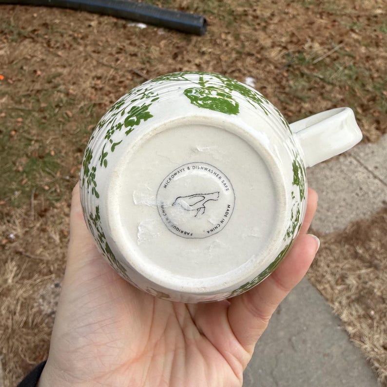 May include: White ceramic mug with a green floral pattern. The mug has a handle and a rounded shape. The bottom of the mug has a circular label with text and a bird illustration. The mug is labeled as microwave and dishwasher safe.