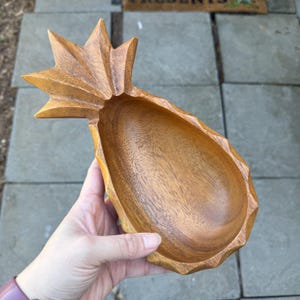May include: A wooden pineapple shaped bowl with a natural wood finish. The bowl is carved to resemble a pineapple with a detailed top and a smooth, rounded interior.