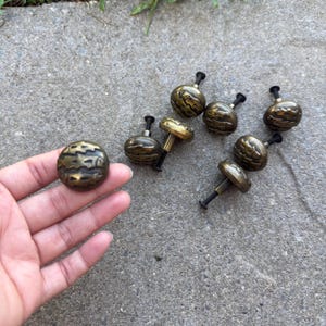 May include: A selection of antique brass cabinet knobs with a textured, ridged pattern. One knob is held in a hand, with others on a concrete surface. The knobs have a dark, aged finish, suggesting vintage or rustic decor. The knobs are likely measured in centimetres.