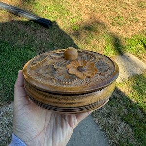 Puede incluir: Una caja redonda de madera con tapa, con diseños florales y de hojas tallados. La tapa tiene una flor prominente y un pequeño pomo. La caja se sostiene en una mano, mostrando su detallada artesanía y tonos de madera natural.