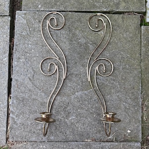 May include: A pair of gold-toned metal wall sconces featuring elongated, curved wire frames with decorative scrollwork at the top and a circular candle holder base at the bottom. The design consists of slender, polished metal rods that form an S-shape.