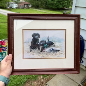 Puede incluir: Impresión enmarcada que presenta tres cachorros de labrador negro con un pato. La obra de arte está ambientada en una escena natural al aire libre con hierba alta y un fondo de color claro. El marco es de color marrón oscuro.