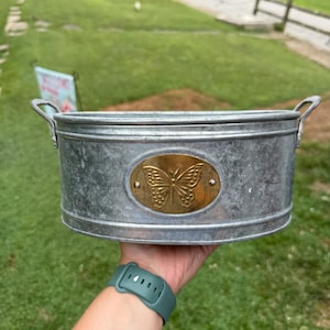 May include: Oval galvanized metal planter with a brass butterfly emblem. The planter has two small handles and is held in a hand against a green background. The planter is approximately 12 inches long.