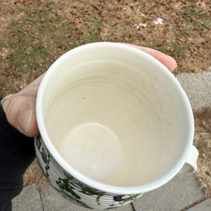 May include: A cream-colored ceramic mug with a ribbed exterior and a green floral pattern. The mug has a small handle and is held in a person's hand. The interior of the mug is smooth and clean.