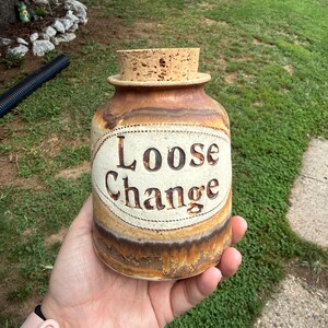 May include: A cylindrical ceramic jar with a cork stopper, featuring a tan and brown drip glaze finish. The front displays an oval cream-colored panel with the words Loose Change embossed in dark brown lettering, creating a textured appearance.