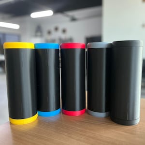 Peut inclure: Cinq contenants cylindriques noirs avec des dessus et des dessous colorés. Les dessus et les dessous sont jaunes, bleus, rouges, gris et noirs. Les contenants sont disposés en rangée sur une surface en bois.