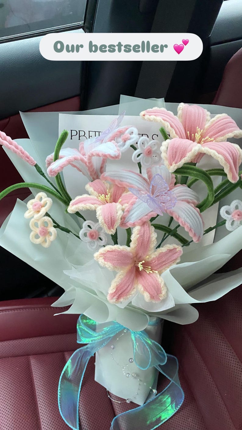 May include: A bouquet of handmade pink and white lilies, accented with green stems and delicate white and lavender butterflies. The bouquet is wrapped in light blue paper and tied with a shimmering blue ribbon. Text reads "Our bestseller".