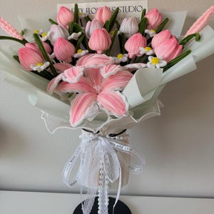 May include: A bouquet of pink and white fabric tulips and a large pink lily, wrapped in light green paper. The bouquet is tied with a white ribbon and a string of pearls. The text "TY FLOWERS STUDIO" is visible on a card.