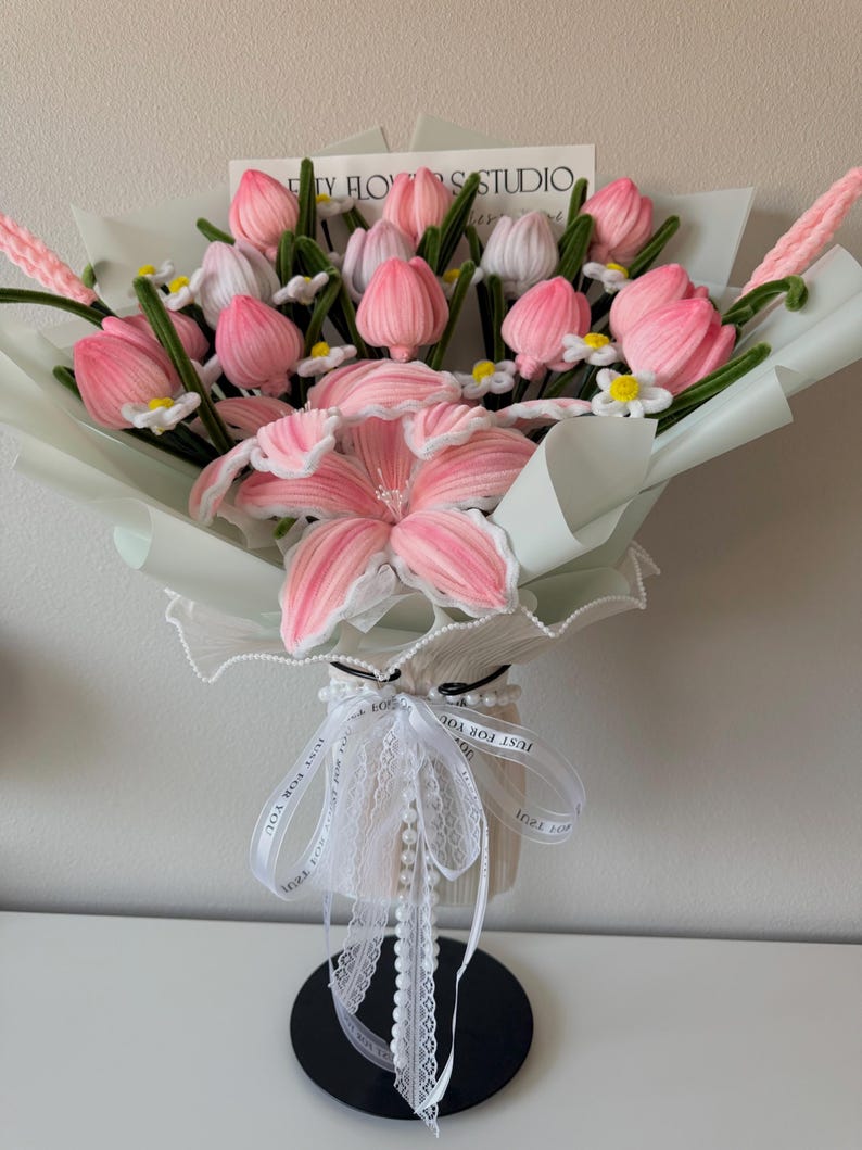 May include: A bouquet of pink and white fabric tulips and a large pink lily, wrapped in light green paper. The bouquet is tied with a white ribbon and a string of pearls. The text "TY FLOWERS STUDIO" is visible on a card.