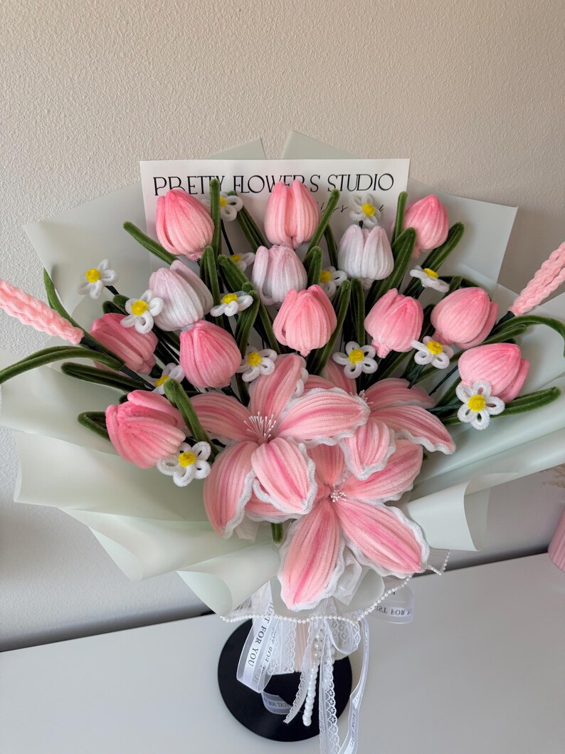 May include: A bouquet of pink and white fabric flowers, including tulips and lilies, wrapped in light green paper. The bouquet is tied with a white ribbon and features the text "PRETTY FLOWERS STUDIO".