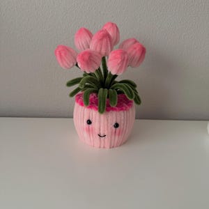 May include: A pink, plush flower pot with a smiling face, holding a bouquet of pink tulips. The tulips have green stems and leaves. The pot is on a white surface.