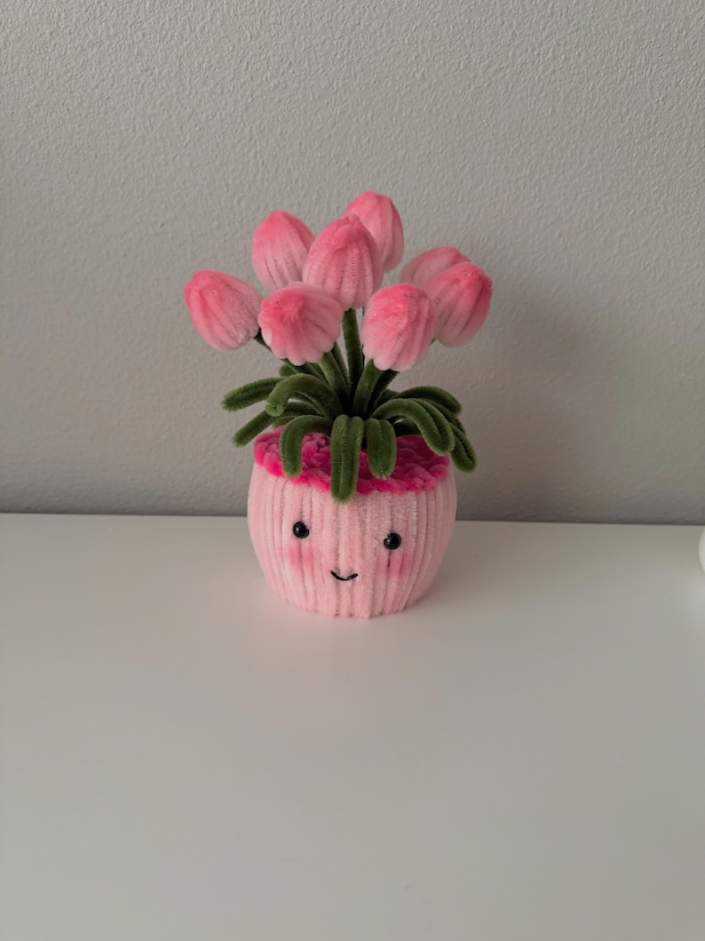 May include: A pink, plush flower pot with a smiling face, holding a bouquet of pink tulips. The tulips have green stems and leaves. The pot is on a white surface.