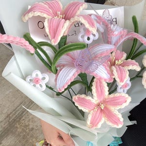 May include: A bouquet of handmade pink and white flowers with green stems and leaves. The flowers are crafted with a soft, textured material, and some have delicate butterfly accents. The bouquet is wrapped in light blue paper.