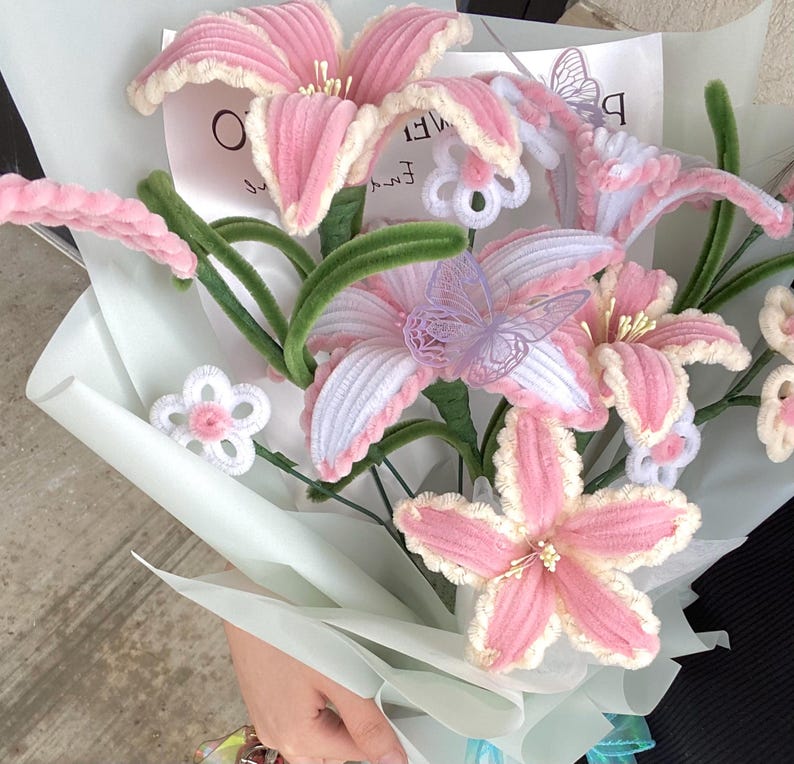 May include: A bouquet of handmade pink and white flowers with green stems and leaves. The flowers are crafted with a soft, textured material, and some have delicate butterfly accents. The bouquet is wrapped in light blue paper.