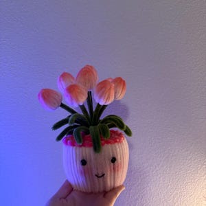 May include: A handmade, whimsical decorative plant in a small, off-white pot with a smiling face. The plant features pink and peach-colored flower buds and green leaves. The pot is held in a hand against a purple-toned background.