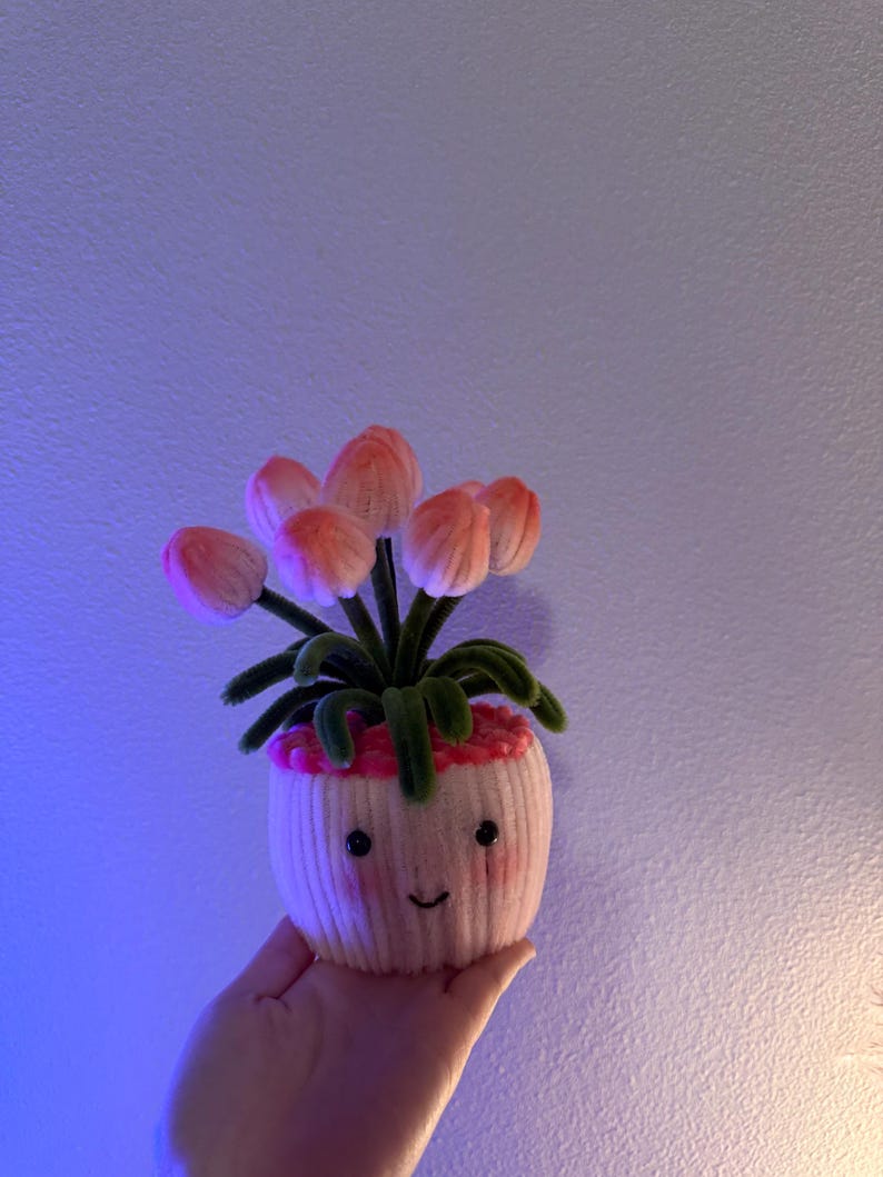 May include: A handmade, whimsical decorative plant in a small, off-white pot with a smiling face. The plant features pink and peach-colored flower buds and green leaves. The pot is held in a hand against a purple-toned background.