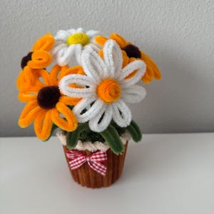 May include: A handcrafted floral arrangement featuring pipe cleaner daisies in a small brown pot. The flowers are white, yellow, and orange with brown centers. A red and white checkered bow adorns the pot, adding a decorative touch.