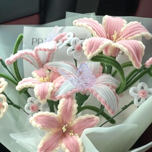 May include: A bouquet of handmade pink and white crochet flowers with green stems and decorative butterflies. The flowers are wrapped in white paper, creating a soft and delicate arrangement.