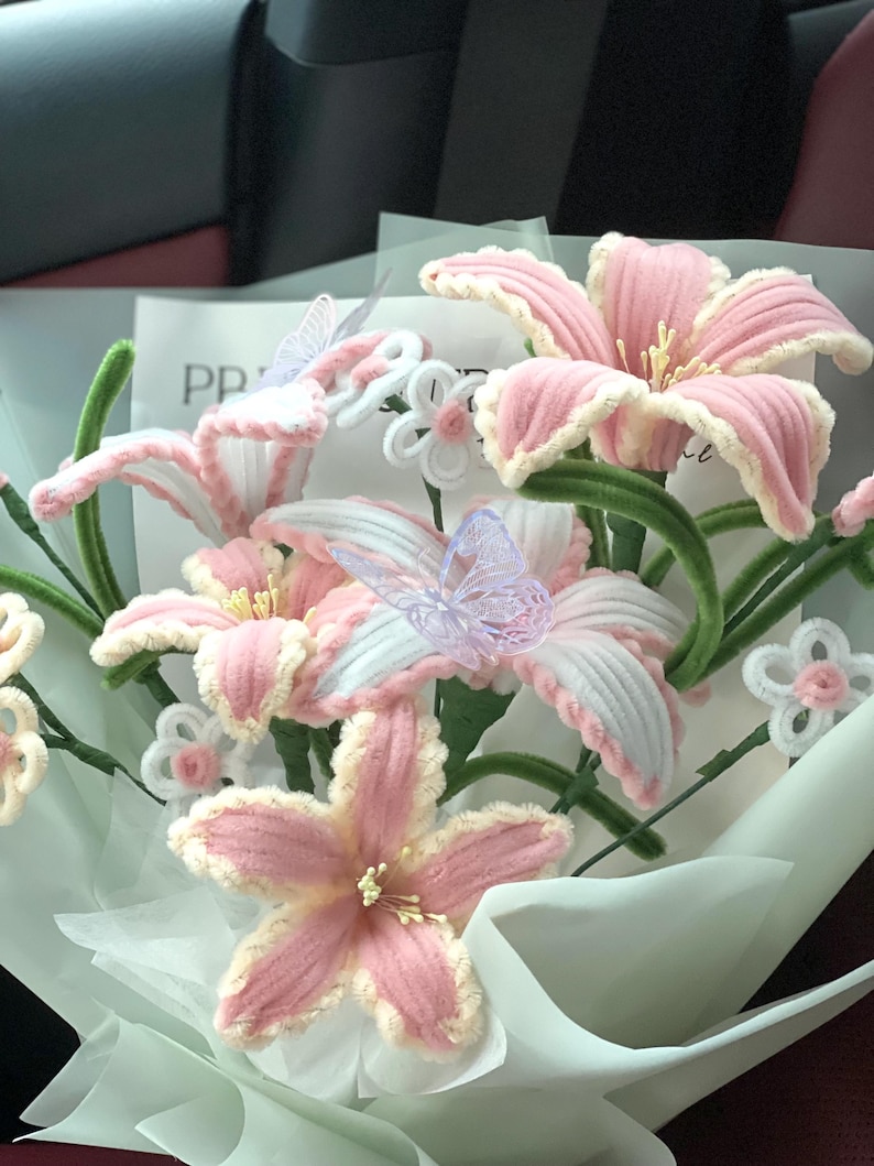 May include: A bouquet of handmade pink and white crochet flowers with green stems and decorative butterflies. The flowers are wrapped in white paper, creating a soft and delicate arrangement.