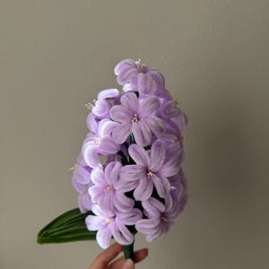 May include: A handcrafted, lavender-coloured hyacinth flower made of soft material. The flower has multiple petals and a green stem with a single green leaf. The flower is held against a neutral background.