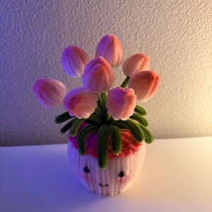 May include: A whimsical handmade flower arrangement featuring pink tulips in a white pot with a smiling face. The pot has a pink rim and green stems and leaves. The background is a gradient of purple and white.