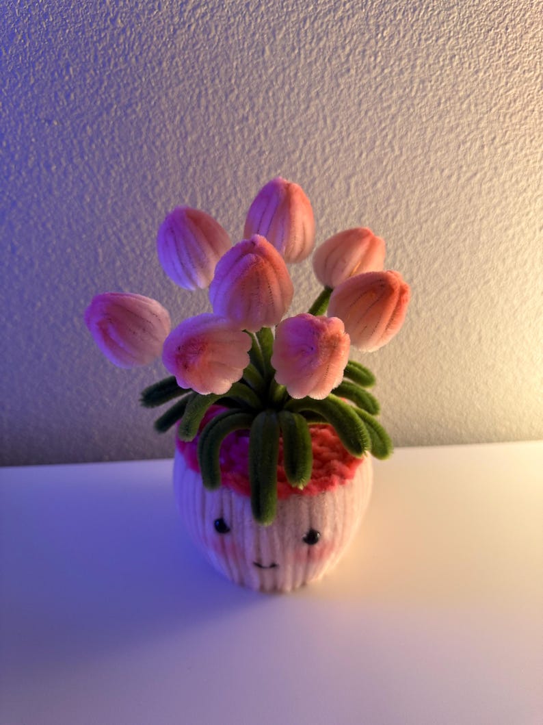 May include: A whimsical handmade flower arrangement featuring pink tulips in a white pot with a smiling face. The pot has a pink rim and green stems and leaves. The background is a gradient of purple and white.