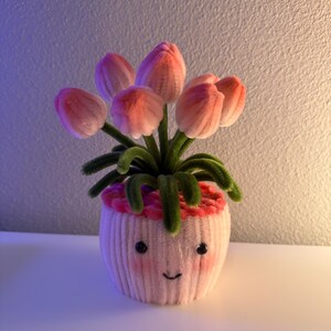 May include: A whimsical, handmade flower pot featuring pink tulips. The pot is a light pink color with a smiling face and rosy cheeks. The tulips have pink petals and green stems, creating a cheerful, decorative piece.