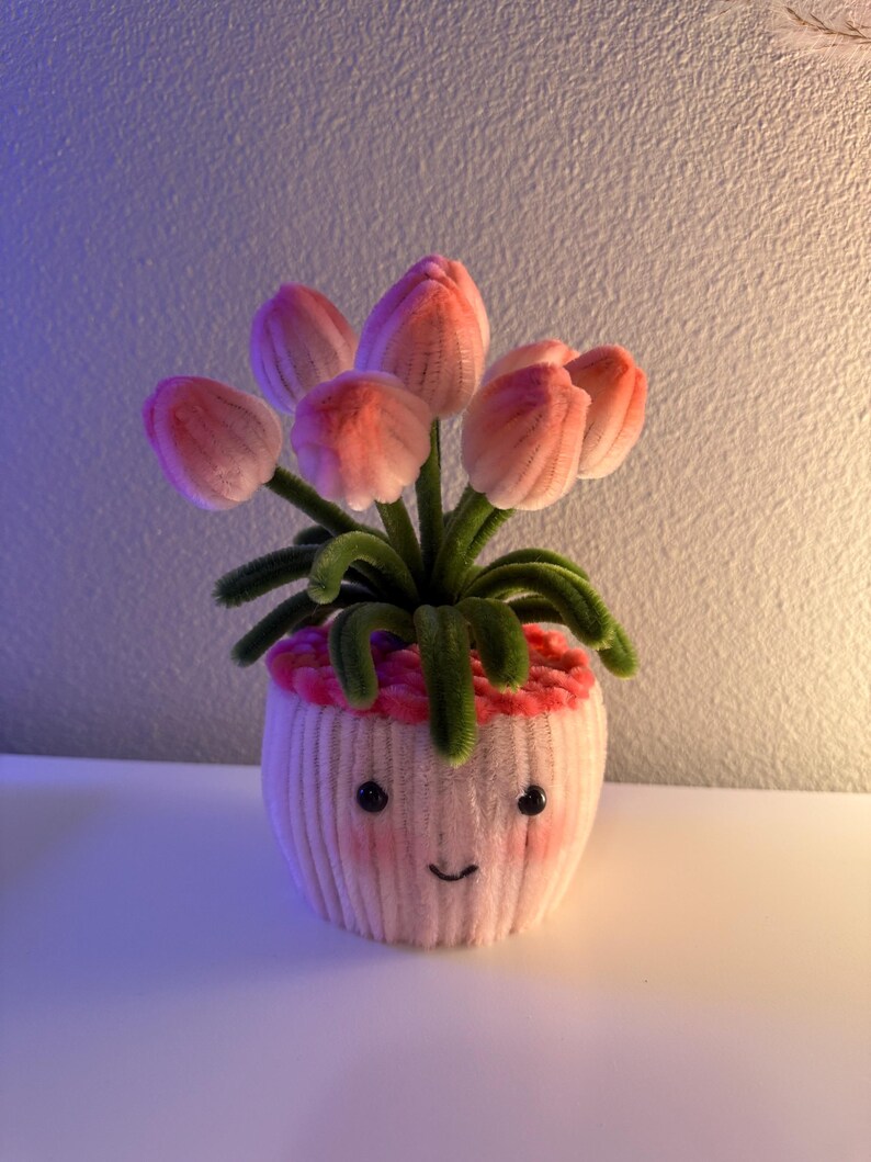 May include: A whimsical, handmade flower pot featuring pink tulips. The pot is a light pink color with a smiling face and rosy cheeks. The tulips have pink petals and green stems, creating a cheerful, decorative piece.