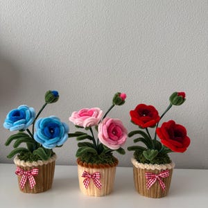 May include: Three decorative flower arrangements in cupcake-shaped pots. The flowers are blue, pink, and red, with green stems and leaves. Each pot is tan with a red and white checkered bow.
