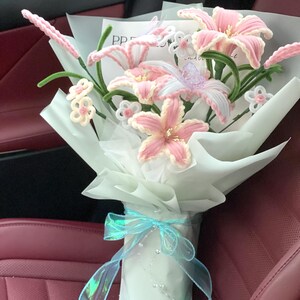 May include: A bouquet of handmade pink and white lilies wrapped in white paper, tied with a light blue ribbon. The lilies have green stems and are accented with small white and pink flowers.