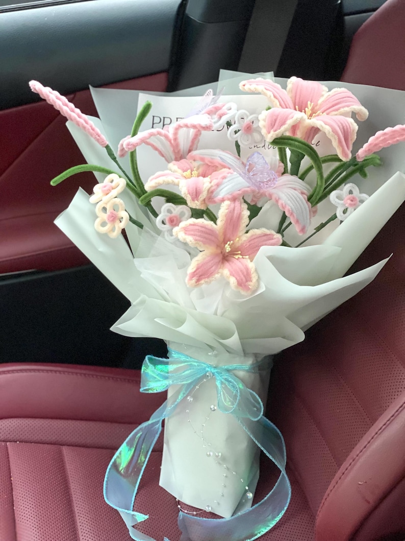 May include: A bouquet of handmade pink and white lilies wrapped in white paper, tied with a light blue ribbon. The lilies have green stems and are accented with small white and pink flowers.
