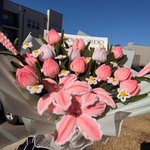 May include: A bouquet of pink and white fabric flowers, including tulips and lilies, wrapped in light green paper. The bouquet is tied with a white ribbon that says "Just For You". The background includes a building and a clear blue sky.