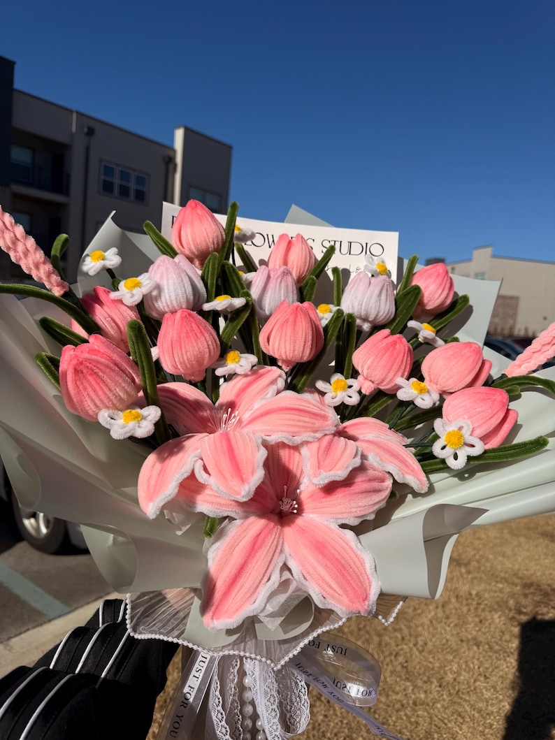 May include: A bouquet of pink and white fabric flowers, including tulips and lilies, wrapped in light green paper. The bouquet is tied with a white ribbon that says "Just For You". The background includes a building and a clear blue sky.