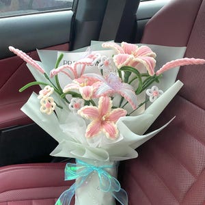 May include: A bouquet of handmade pink and white crochet lilies and other flowers, wrapped in light blue paper and tied with a blue iridescent ribbon. The bouquet is in a car with red seats.