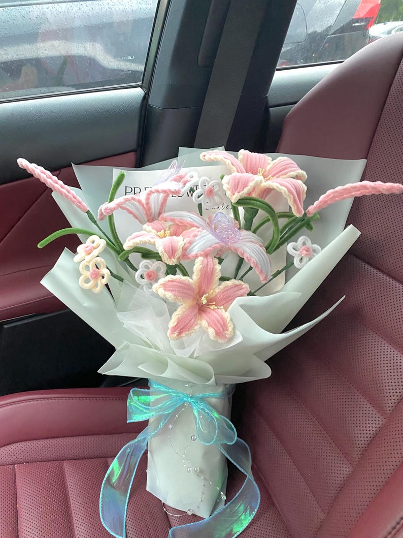 May include: A bouquet of handmade pink and white crochet lilies and other flowers, wrapped in light blue paper and tied with a blue iridescent ribbon. The bouquet is in a car with red seats.