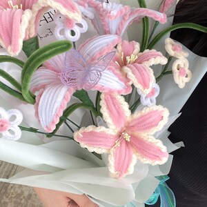 May include: A bouquet of handmade pink and white flowers with green stems and decorative butterflies. The flowers are wrapped in light blue paper and tied with a blue ribbon. The flowers are made of a soft, textured material.
