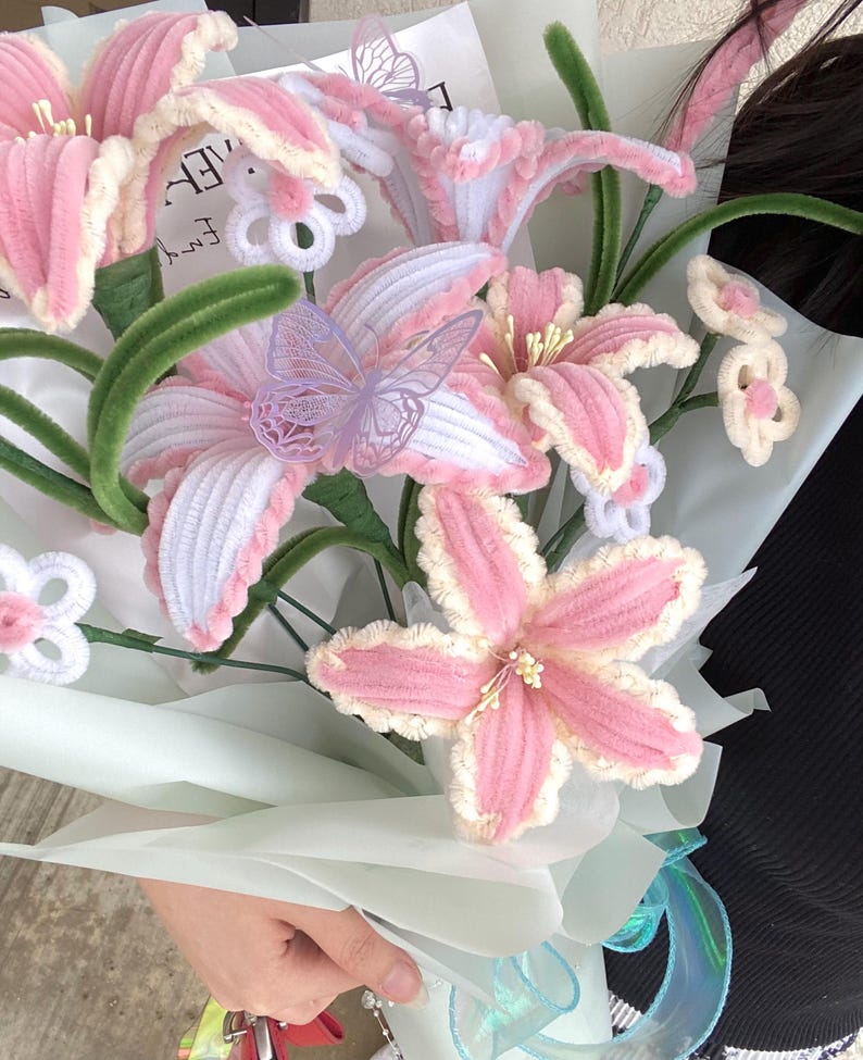 May include: A bouquet of handmade pink and white flowers with green stems and decorative butterflies. The flowers are wrapped in light blue paper and tied with a blue ribbon. The flowers are made of a soft, textured material.