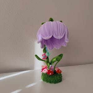 May include: A handcrafted pipe cleaner flower sculpture featuring a large, light purple bell-shaped bloom atop a green stem. The base is covered in green moss and decorated with small red and white mushrooms, tiny pink and blue flowers, and green leaves.