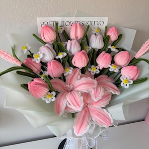 May include: A bouquet of pink and white fabric flowers, including tulips and lilies, wrapped in light green paper. The bouquet is tied with a white ribbon and features the text "PRETTY FLOWERS STUDIO".