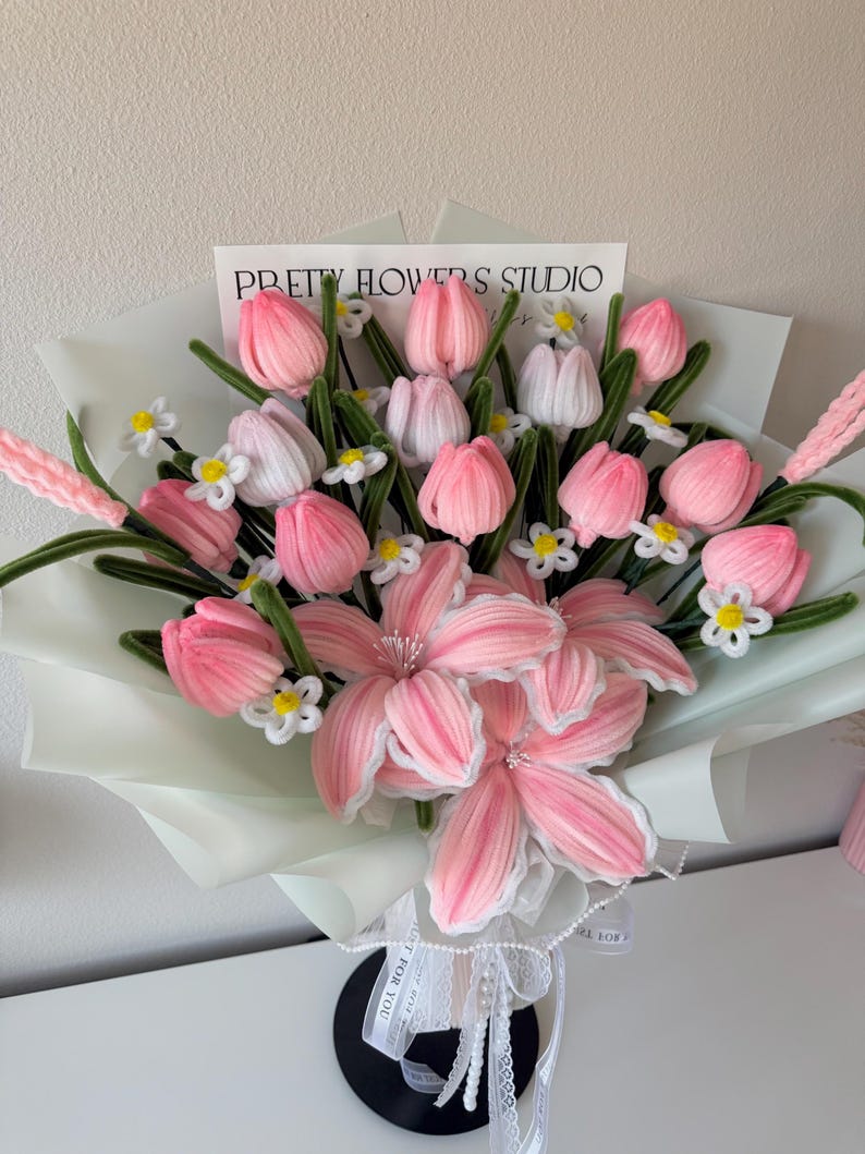 May include: A bouquet of pink and white fabric flowers, including tulips and lilies, wrapped in light green paper. The bouquet is tied with a white ribbon and features the text "PRETTY FLOWERS STUDIO".