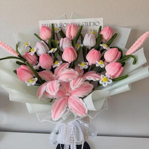 May include: A bouquet of pink and white fabric flowers, including tulips and lilies, wrapped in light green paper. The bouquet is tied with a white ribbon that says "JUST FOR YOU". A card that says "PRETTY FLOWERS STUDIO" is visible.