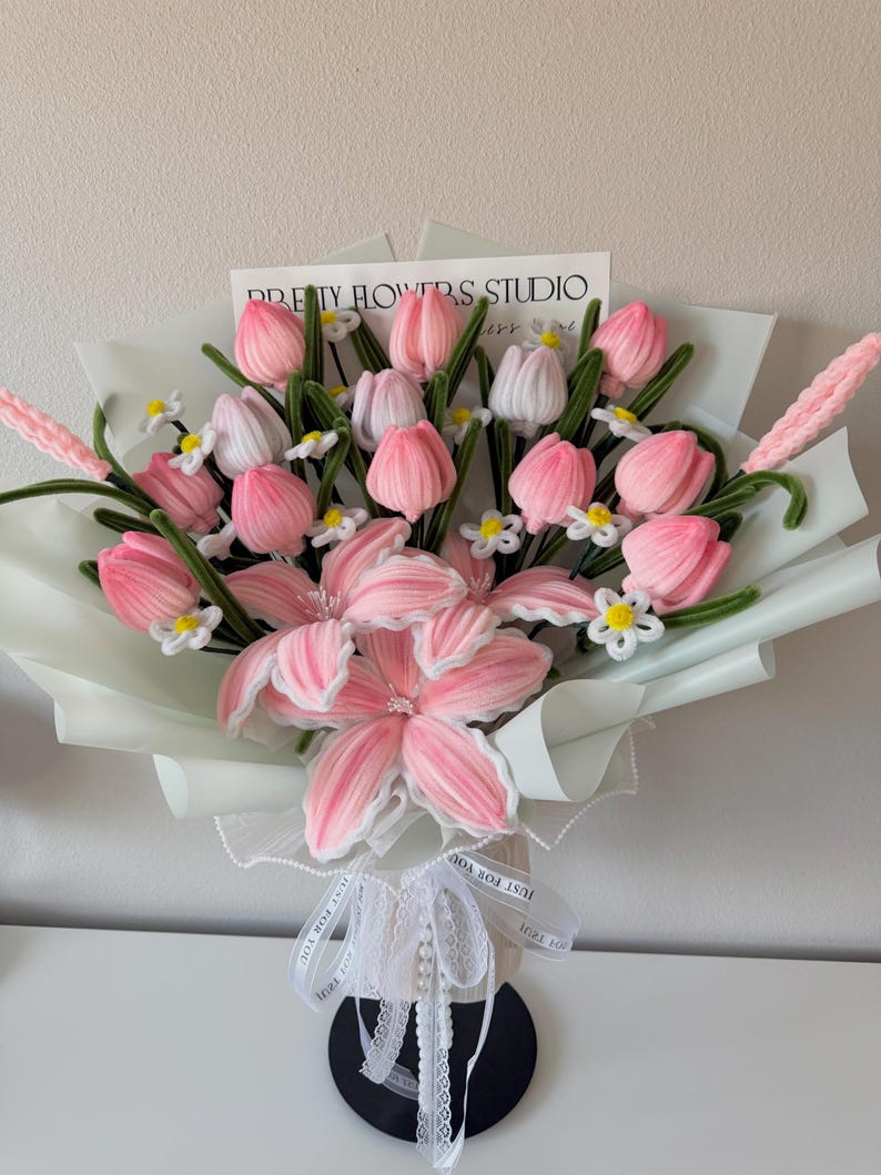 May include: A bouquet of pink and white fabric flowers, including tulips and lilies, wrapped in light green paper. The bouquet is tied with a white ribbon that says "JUST FOR YOU". A card that says "PRETTY FLOWERS STUDIO" is visible.