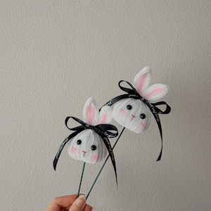 May include: Two white bunny decorations with pink inner ears, black eyes, and a small red "T" for a mouth. Each bunny has a black ribbon tied in a bow with the words "for you" printed on it. The bunnies are attached to green wire stems.