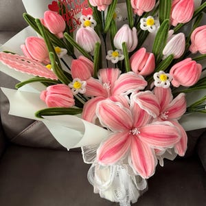 May include: A bouquet of pink and white flowers, including tulips and lilies, wrapped in white paper. The bouquet has green stems and small white flowers. A red heart-shaped decoration with the words "Happy Valentine's Day" is visible.