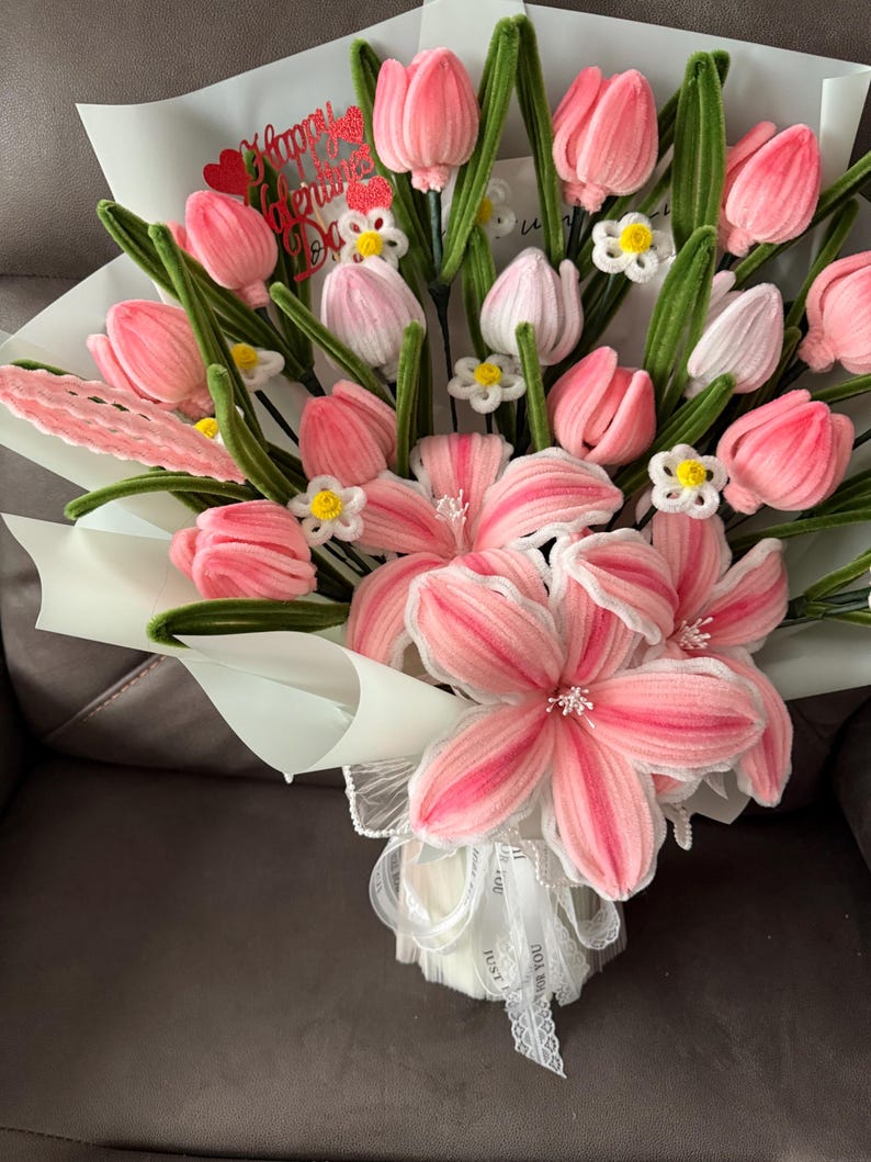 May include: A bouquet of pink and white flowers, including tulips and lilies, wrapped in white paper. The bouquet has green stems and small white flowers. A red heart-shaped decoration with the words "Happy Valentine's Day" is visible.