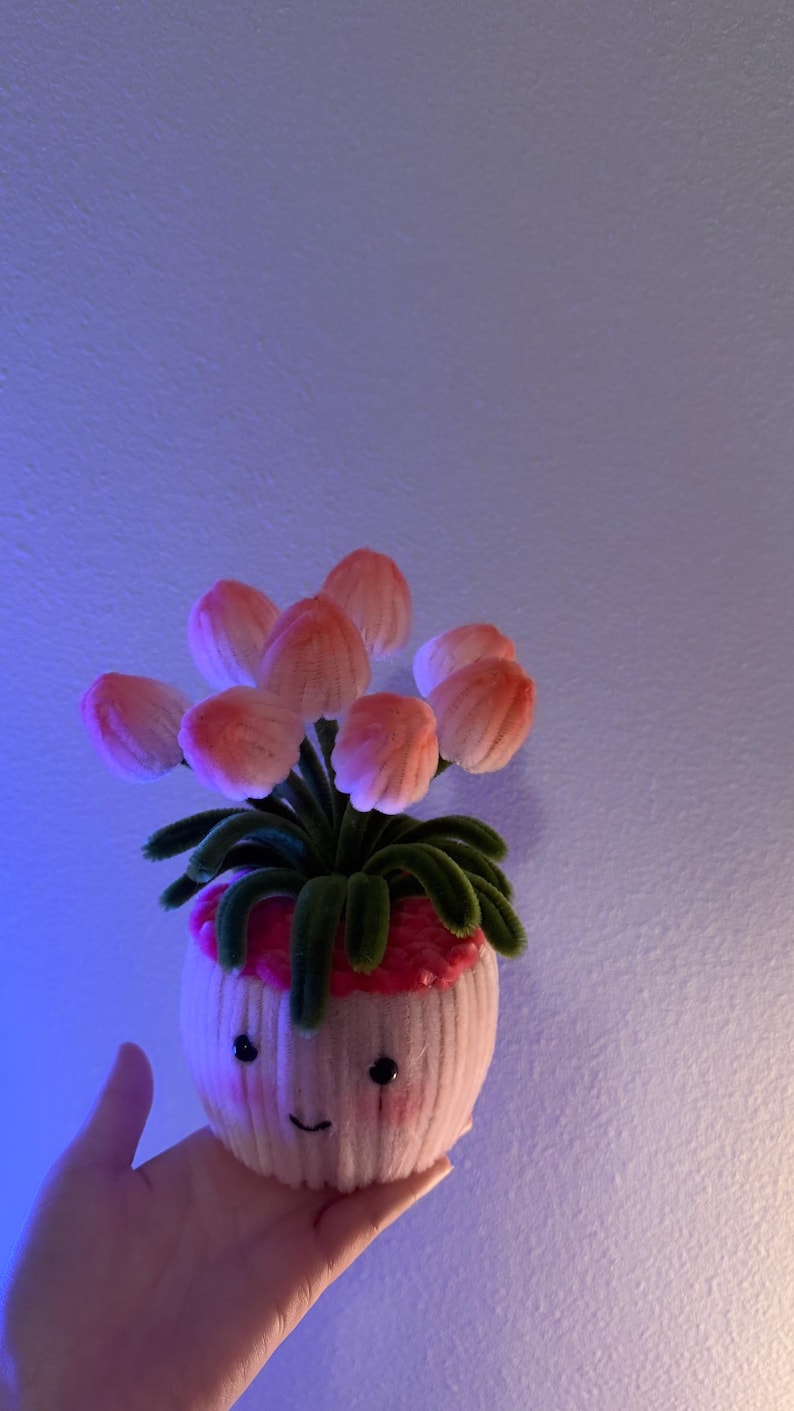 May include: A whimsical, handmade felt flower pot with a smiling face. The pot features pink and white felt, with green felt leaves and pink flower petals. The craft item is a unique decorative piece.