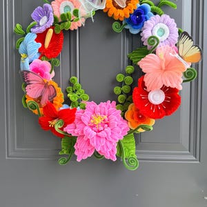 May include: A circular wreath constructed from colorful pipe cleaners shaped into various flowers, including a large central pink bloom, red hibiscus, and orange blossoms. The design features green spiraled stems, leaves, and two butterfly cutouts.