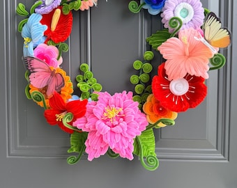 May include: A circular wreath constructed from colorful pipe cleaners shaped into various flowers, including a large central pink bloom, red hibiscus, and orange blossoms. The design features green spiraled stems, leaves, and two butterfly cutouts.